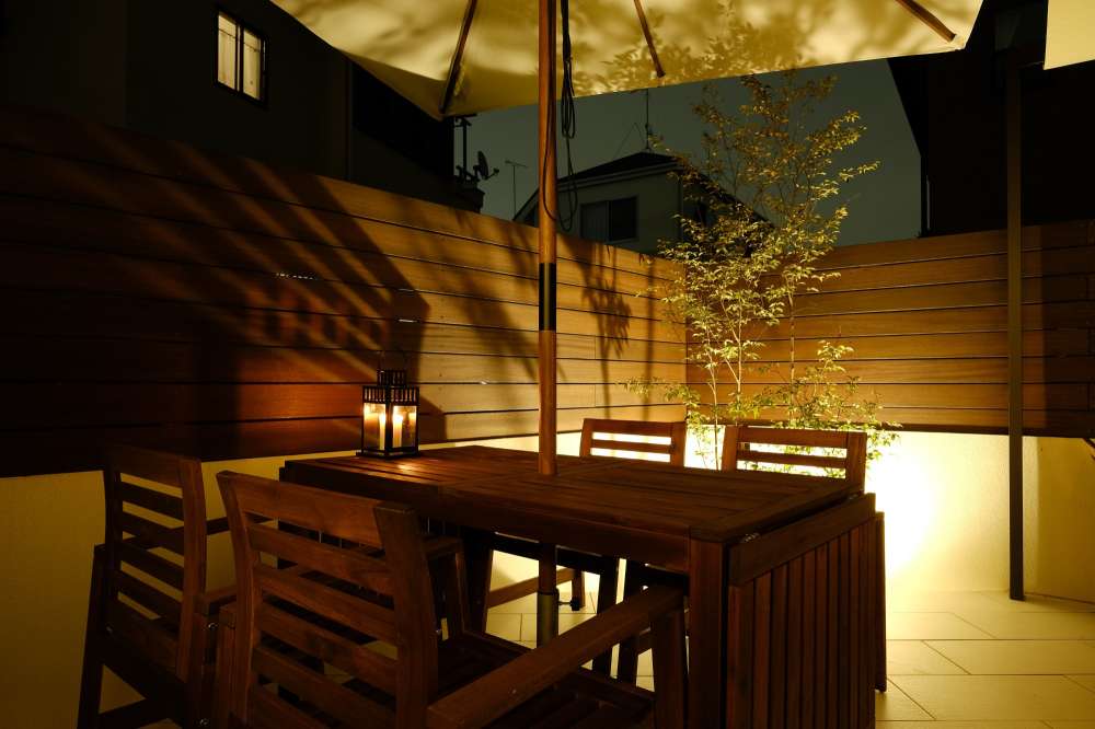 ウッドテーブル越しに見えるウッドフェンスと樹木を照らすスポットライトの光